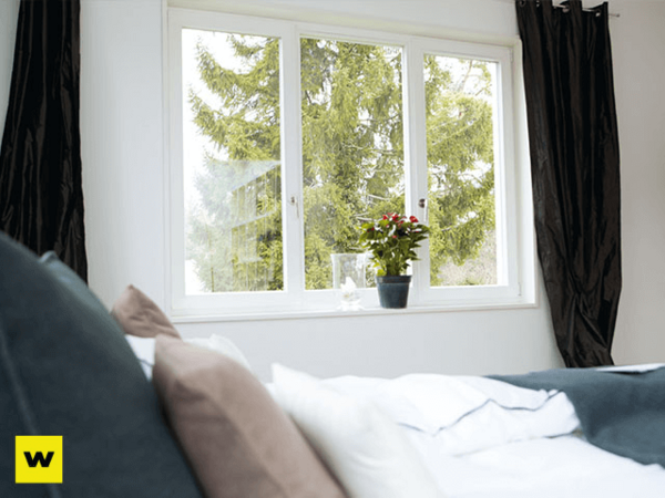 Helles Schlafzimmer mit großem Fenster, Blick ins Grüne, Bett und Zimmerpflanze auf der Fensterbank.